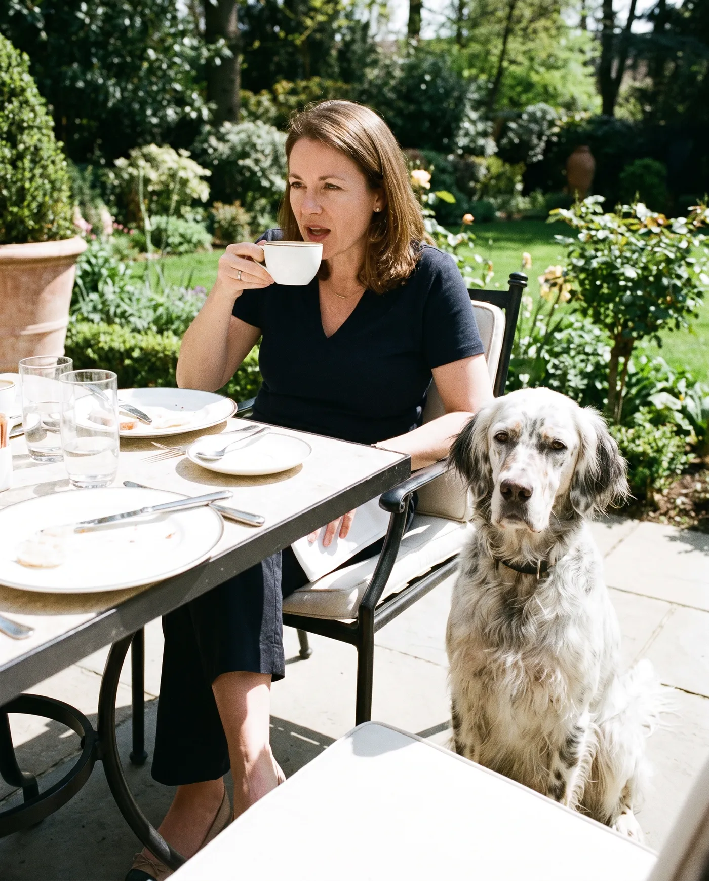 A woman drinking coffee at a garden table with an English setter sitting attentively at her side