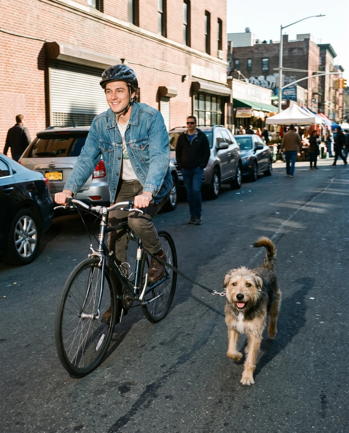 A man cycling down a city street with a tan dog running steadily beside the bike