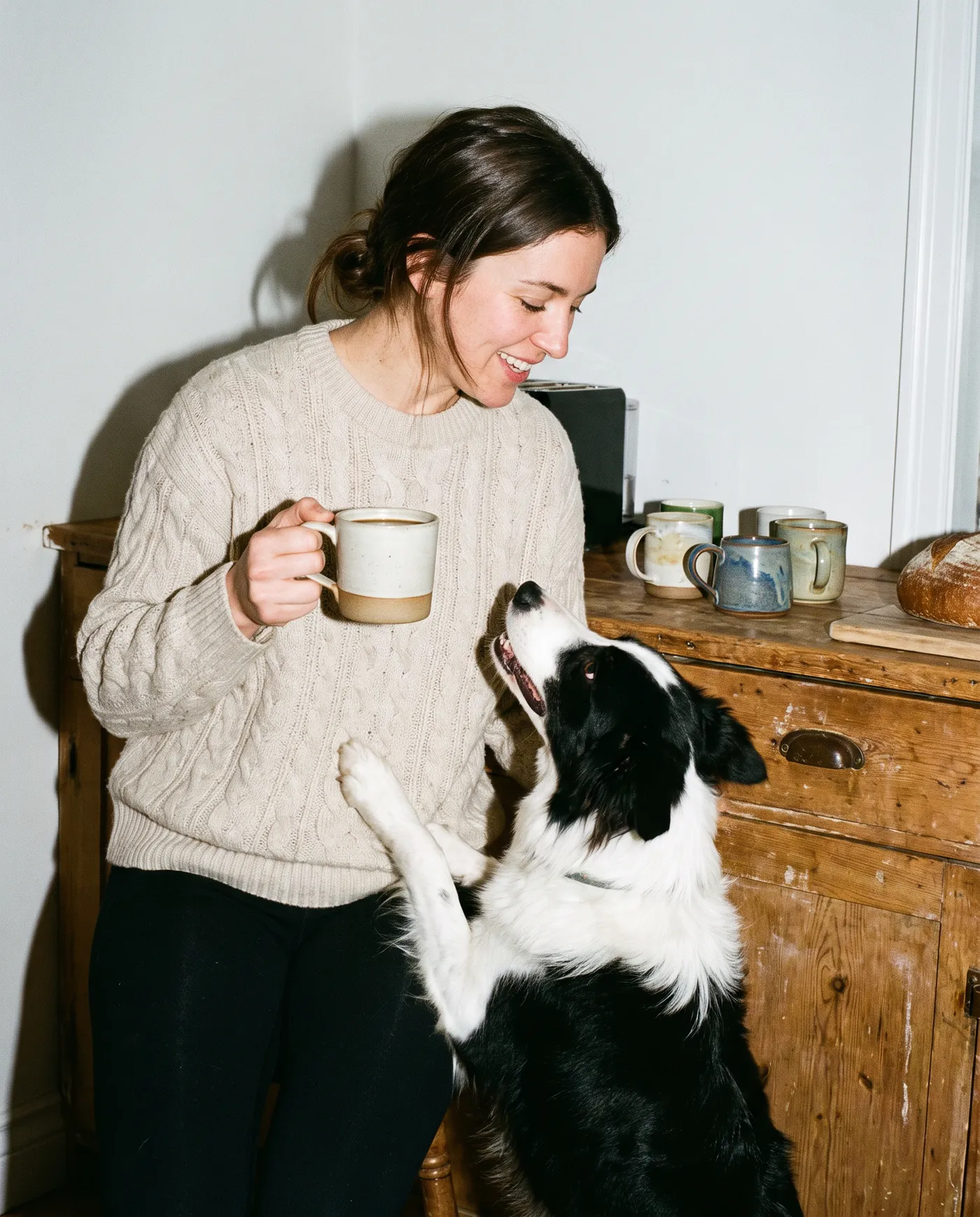 A woman holding a mug in a farmhouse kitchen as her black and white collie jumps up to greet her