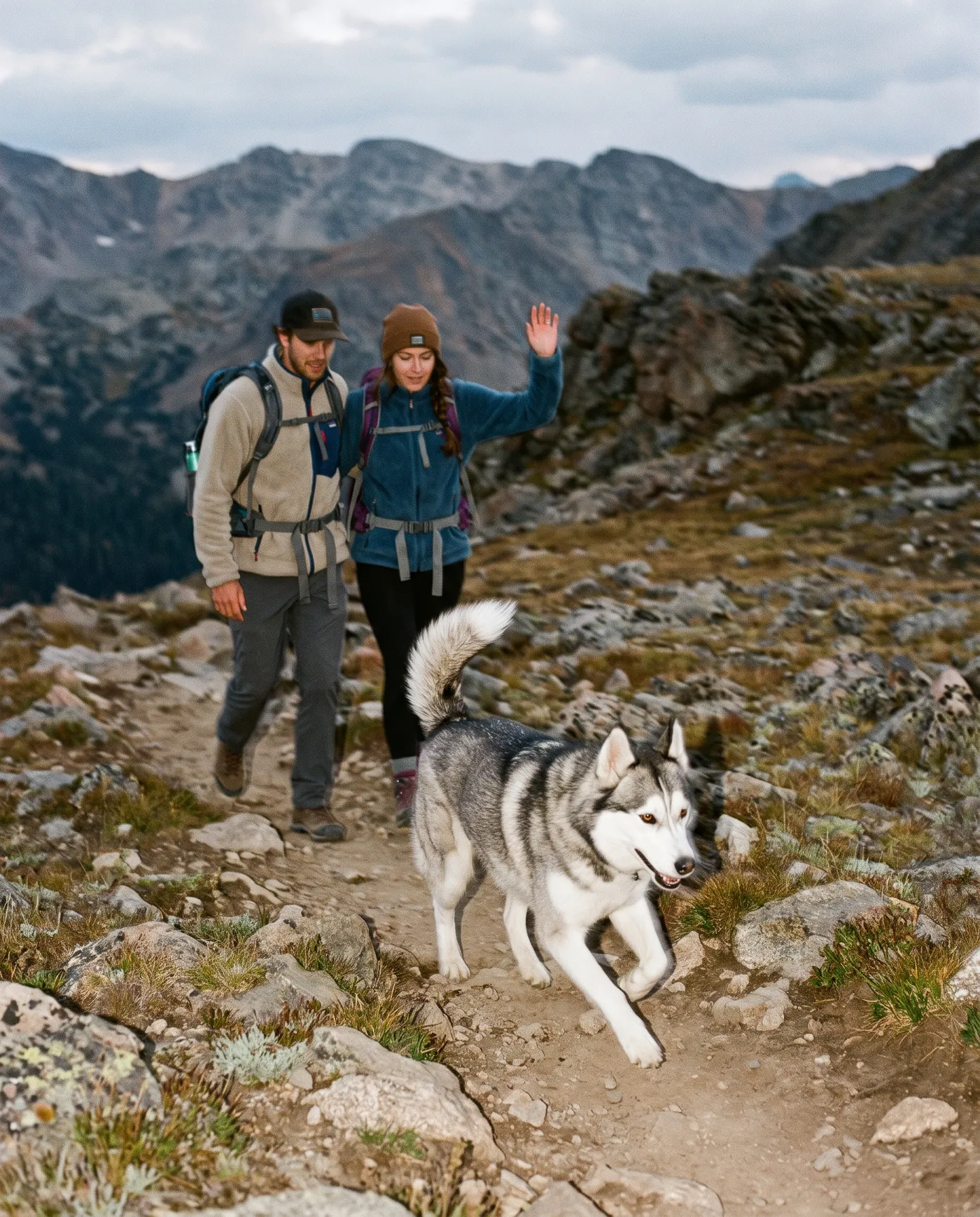 Two hikers with backpacks crossing a rocky mountain ridge with a husky picking its way over the stones