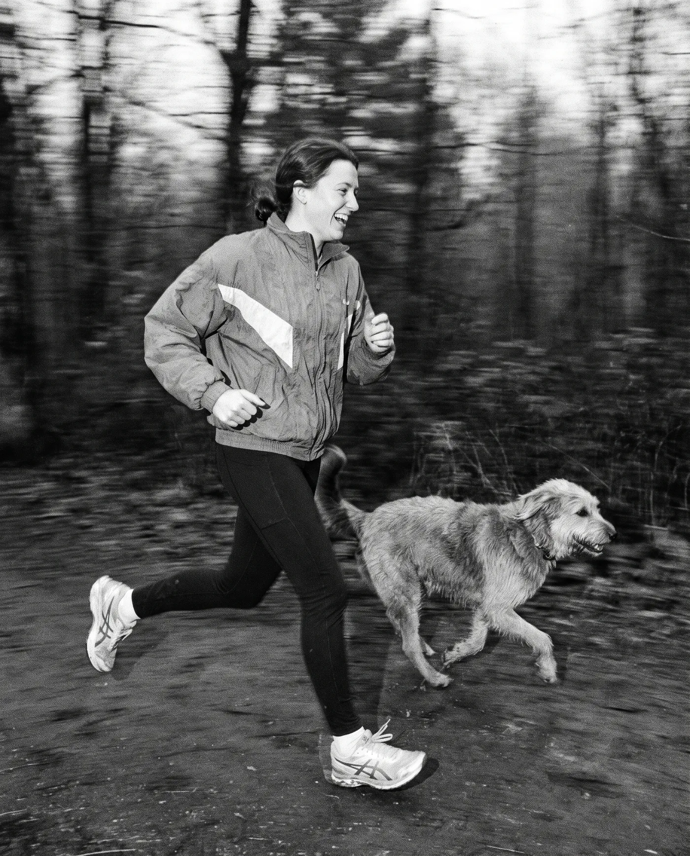 Black and white photograph of a woman jogging along a woodland path with a golden retriever running at her heel